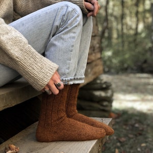 May include: A pair of brown knitted socks with a ribbed texture. The socks are worn by a person sitting on wooden steps with fall leaves scattered around.