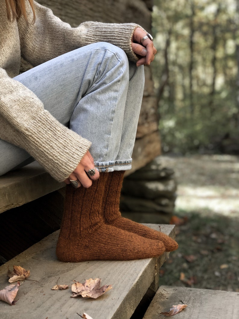 May include: A pair of brown knitted socks with a ribbed texture. The socks are worn by a person sitting on wooden steps with fall leaves scattered around.