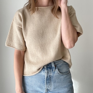 May include: A beige knitted short-sleeved top with a relaxed fit. The top is worn with light blue denim jeans.