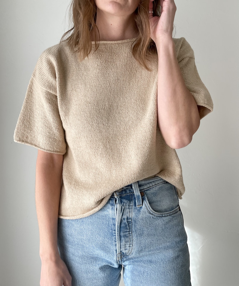 May include: A beige knitted short-sleeved top with a relaxed fit. The top is worn with light blue denim jeans.