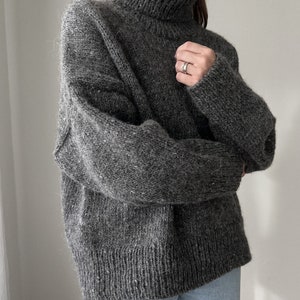 Puede incluir: Un su&eacute;ter gris de cuello alto de gran tama&ntilde;o con un corte holgado. El su&eacute;ter est&aacute; hecho de un hilo suave y peludo.
