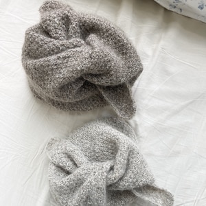 May include: Two knitted beanies in shades of gray and taupe, with a knotted design on top. The beanies are made of a textured yarn and are displayed on a white surface. The hats appear soft and warm, suitable for cold weather.