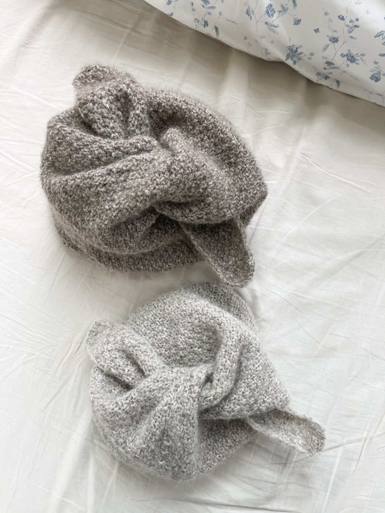 May include: Two knitted beanies in shades of gray and taupe, with a knotted design on top. The beanies are made of a textured yarn and are displayed on a white surface. The hats appear soft and warm, suitable for cold weather.