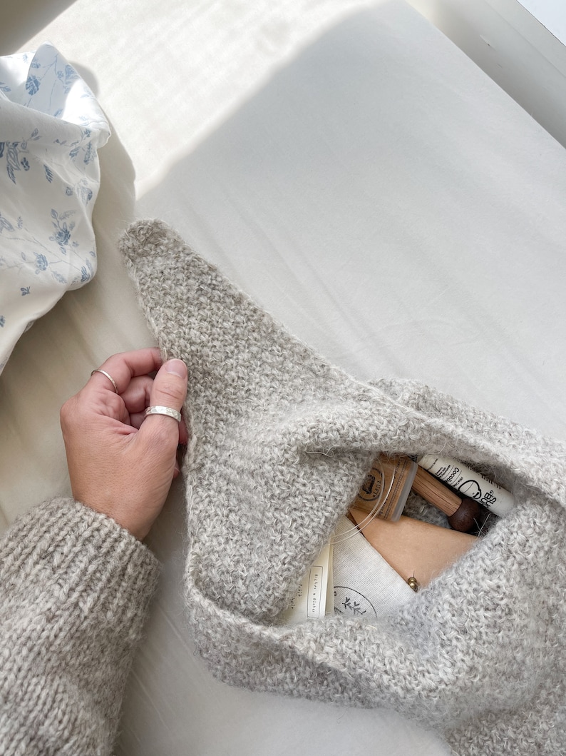 May include: A light gray knitted bag, partially open, revealing small items inside. The bag is held by a hand with a silver ring. The items include a small wooden stamp, a small pouch, and other small items. The bag is on a white surface.