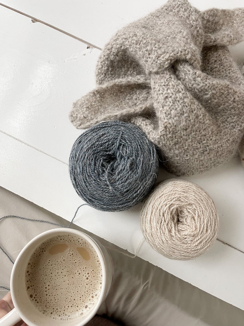 May include: A cozy scene featuring a mug of coffee with a light brown beverage, two balls of yarn in gray and beige, and a knitted scarf. The yarn is ready for knitting or crocheting projects.