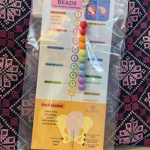 May include: A set of dilation beads in a clear plastic package. The beads are in a rainbow of colors, each representing a centimeter of dilation. The card includes text about labor stages and fetal station.