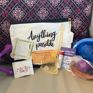 May include: A selection of self-care products, featuring a white canvas bag with the text "Anything is possible", a purple massage tool, and a blue basin. Other items include a comb, essential oil towelettes, and a "hello baby!" packet.