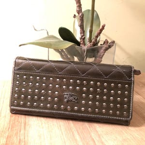 Op de afbeelding: Een donkergroene leren portemonnee met een gestikt patroon en zilveren studs. De portemonnee heeft een Burberry-logo op de voorkant.