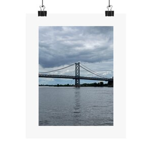Peut inclure: Photographie d'un pont suspendu au-dessus de l'eau sous un ciel nuageux. Le pont est gris et possède des tours de soutien. L'eau est calme et reflète le ciel. L'image est imprimée sur une affiche blanche.