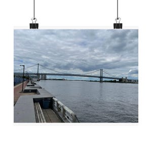 Peut inclure: Impression d'un pont suspendu enjambant une large rivière sous un ciel nuageux. L'image présente une promenade en béton avec une poubelle et une balustrade métallique au premier plan.