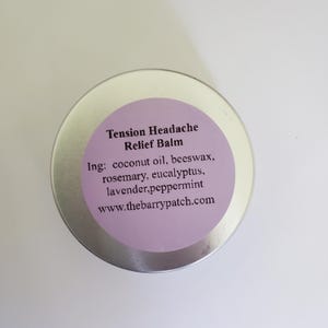 May include: A round, silver-colored tin with a light purple label. The label reads "Tension Headache Relief Balm" and lists ingredients like coconut oil, beeswax, and peppermint. The website www.thebarrypatch.com is also printed on the label.