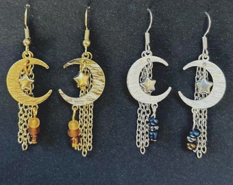 May include: Three pairs of crescent moon earrings with dangling chains and star charms. The earrings are gold, silver, and silver with black beads.