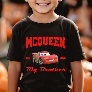 May include: Black t-shirt featuring the name "McQueen" in red block letters above a cartoon image of a red race car. The text "EST 2024" and "Big Brother" are also in red. The shirt is worn by a child.