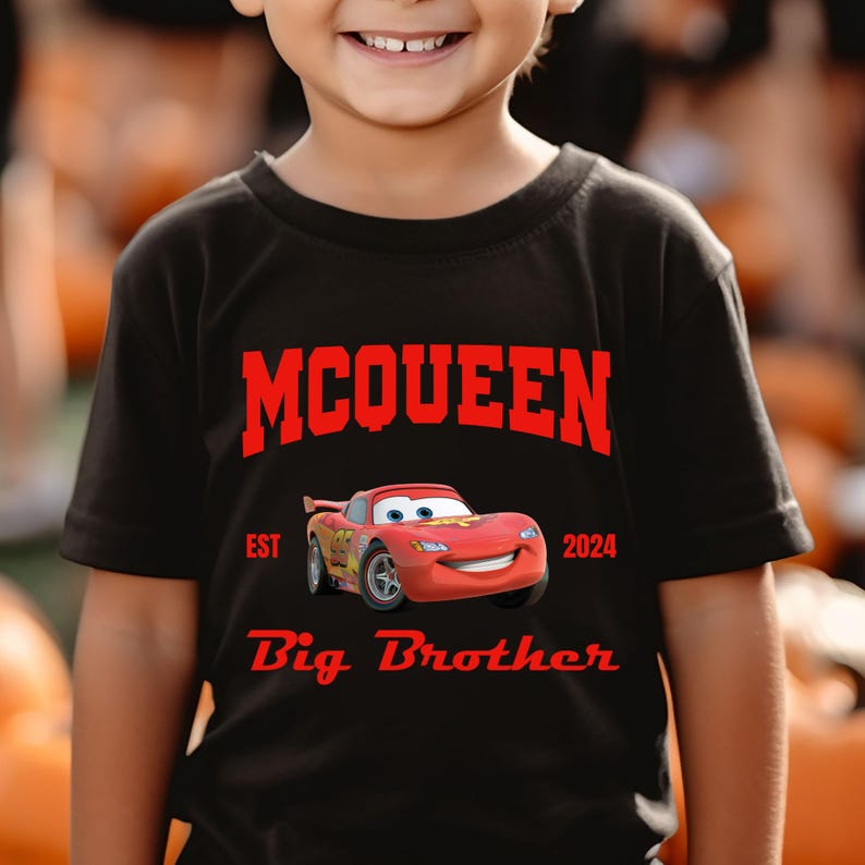 May include: Black t-shirt featuring the name "McQueen" in red block letters above a cartoon image of a red race car. The text "EST 2024" and "Big Brother" are also in red. The shirt is worn by a child.