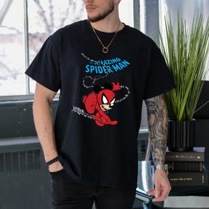 May include: A black short-sleeved t-shirt featuring a graphic of a character with Mickey Mouse ears wearing a red and blue Spider-Man suit. The text THE AMAZING SPIDER-MAN is printed in light blue above the character, who is shown swinging on a web.