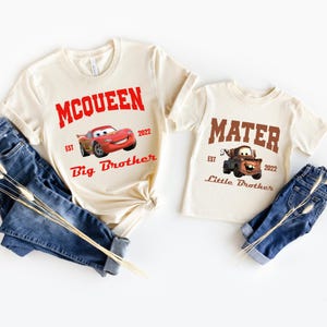 May include: Two beige t-shirts with Cars characters. The larger shirt says "McQueen Est. 2022 Big Brother" with a red Lightning McQueen car. The smaller shirt says "Mater Est. 2022 Little Brother" with a brown Mater tow truck.