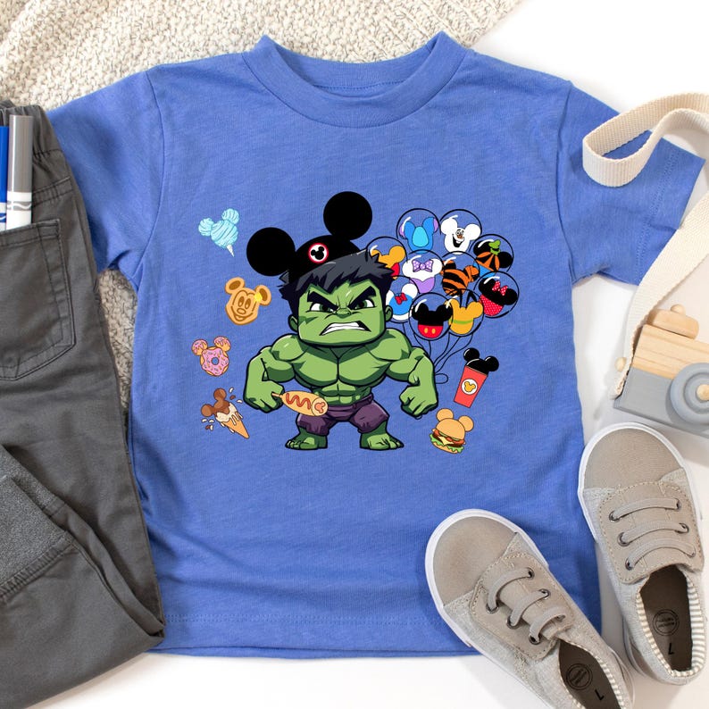 Puede incluir: Una camiseta azul con un personaje verde de Hulk que lleva orejas de Mickey Mouse. Hulk sostiene un perrito caliente y est&aacute; rodeado de globos y bocadillos con tem&aacute;tica de Mickey Mouse.