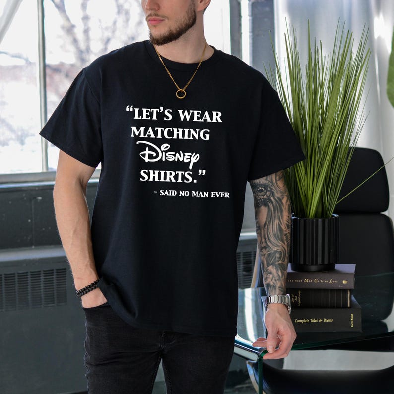 Peut inclure: T-shirt noir avec l'inscription blanche : "LET'S WEAR MATCHING Disney SHIRTS." et "- SAID NO MAN EVER". Le t-shirt est de style col rond et est port&eacute; par une personne.