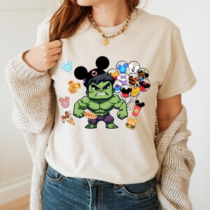 Puede incluir: Una camiseta beige con un personaje verde de Hulk que lleva un sombrero de Mickey Mouse y sostiene un perrito caliente. Hulk est&aacute; rodeado de personajes de dibujos animados y alimentos, incluyendo orejas de Mickey Mouse, una dona, un perrito caliente y una hamburguesa.