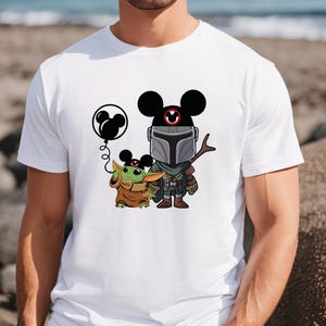 May include: White t-shirt with a cartoon image of a Mandalorian character wearing Mickey Mouse ears and holding a staff.  The Mandalorian is standing next to a small green character wearing Mickey Mouse ears and holding a balloon.