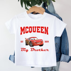 May include: White t-shirt featuring a red graphic of Lightning McQueen from the Cars movie. The shirt includes the text "MCQUEEN", "EST 2024", and "Big Brother" in red. The shirt is hanging on a wooden hanger.