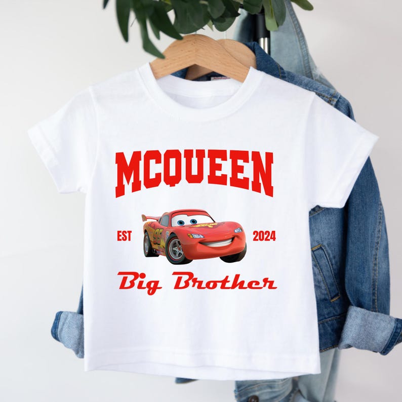 May include: White t-shirt featuring a red graphic of Lightning McQueen from the Cars movie. The shirt includes the text "MCQUEEN", "EST 2024", and "Big Brother" in red. The shirt is hanging on a wooden hanger.