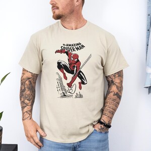 May include: A beige t-shirt with a graphic of Spider-Man swinging through the city. The text "The Amazing Spider-Man" is printed above the graphic.