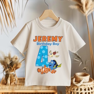 May include: A white t-shirt with an orange and blue design featuring the characters Dory, Nemo, and a turtle. The number 4 is in the center of the design with blue polka dots. The text "JEREMY Birthday Boy" is at the top of the design.