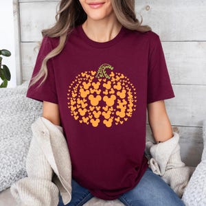 Puede incluir: Camiseta burdeos con un dise&ntilde;o de calabaza hecho de siluetas naranjas de Mickey Mouse. La calabaza tiene un tallo verde. La camiseta es de cuello redondo y manga corta.