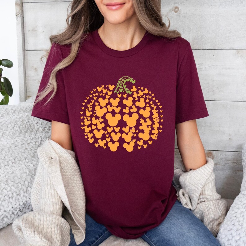 Puede incluir: Camiseta burdeos con un dise&ntilde;o de calabaza hecho de siluetas naranjas de Mickey Mouse. La calabaza tiene un tallo verde. La camiseta es de cuello redondo y manga corta.