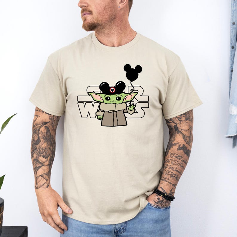 May include: A beige t-shirt featuring a cartoon image of a green alien wearing a Mickey Mouse hat and holding a black balloon. The text "STAR WARS" is printed in black below the image.