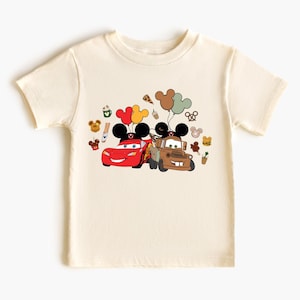May include: Cream-coloured t-shirt featuring a graphic of Lightning McQueen and Mater from the Cars movie, each with Mickey Mouse ears. Balloons and other Disney-themed icons surround the cars. The shirt is made of a soft material.