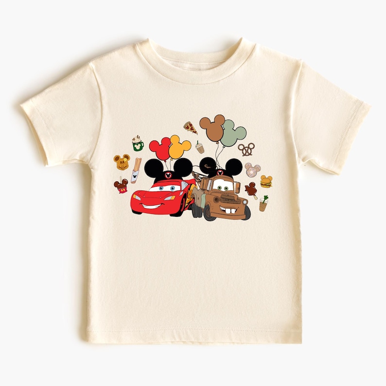 May include: Cream-coloured t-shirt featuring a graphic of Lightning McQueen and Mater from the Cars movie, each with Mickey Mouse ears. Balloons and other Disney-themed icons surround the cars. The shirt is made of a soft material.