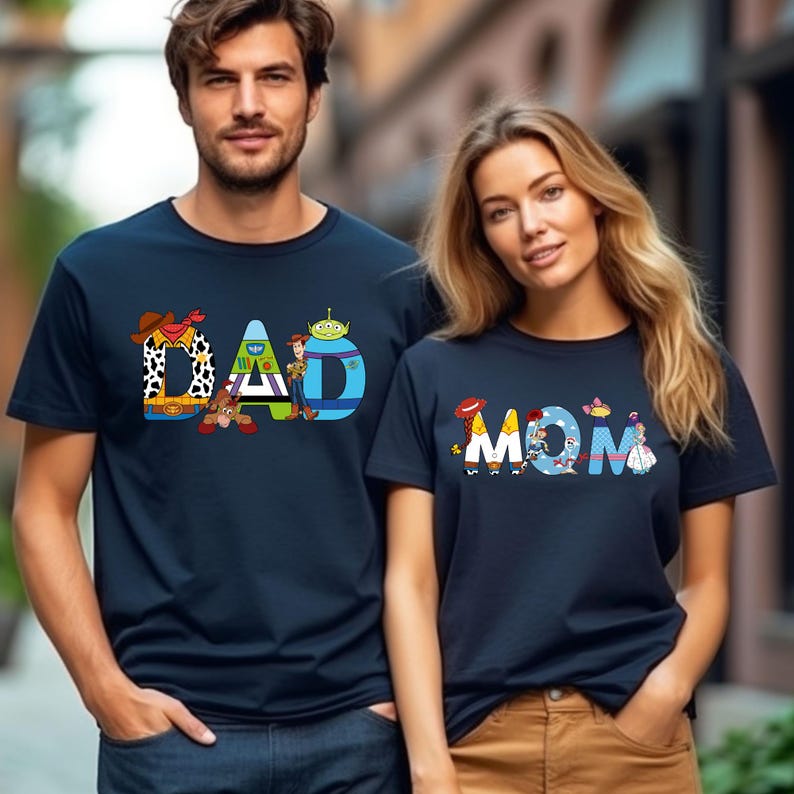 K&ouml;nnte beinhalten: Zwei marineblaue T-Shirts mit den W&ouml;rtern "DAD" und "MOM", die mit Figuren aus dem Toy Story-Film-Franchise geschrieben sind. Die Figuren sind in ihren charakteristischen Posen und Outfits.