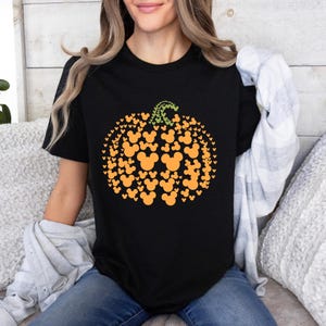 Puede incluir: Camiseta negra con un dise&ntilde;o de calabaza hecho de siluetas naranjas de Mickey Mouse. La calabaza tiene un tallo verde. La camiseta es de cuello redondo. La camiseta se combina con unos vaqueros azules.
