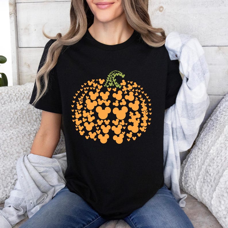 Puede incluir: Camiseta negra con un dise&ntilde;o de calabaza hecho de siluetas naranjas de Mickey Mouse. La calabaza tiene un tallo verde. La camiseta es de cuello redondo. La camiseta se combina con unos vaqueros azules.