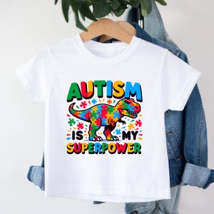Puede incluir: Camiseta blanca con un gr&aacute;fico de dinosaurio colorido y el texto "El autismo es mi superpoder".