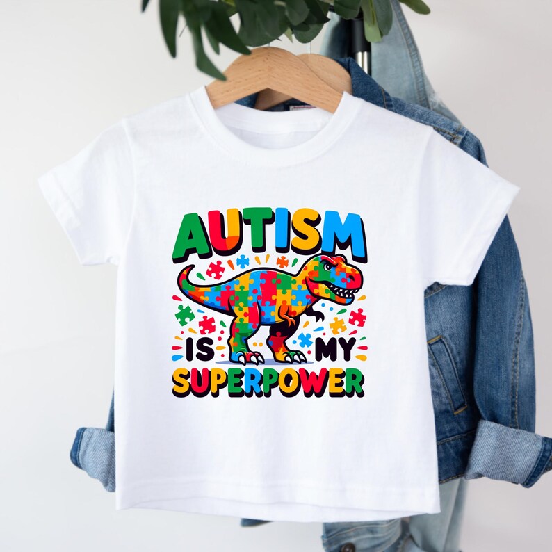 Puede incluir: Camiseta blanca con un gr&aacute;fico de dinosaurio colorido y el texto "El autismo es mi superpoder".