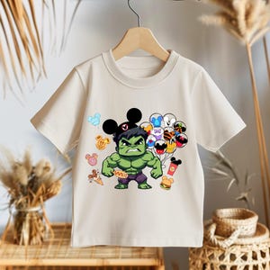 Puede incluir: Una camiseta blanca con un Hulk de dibujos animados verde que lleva un sombrero de Mickey Mouse y sostiene globos. Los globos son de varios colores y tienen orejas de Mickey Mouse. La camiseta tambi&eacute;n tiene un cono de helado de dibujos animados, una dona y un perrito caliente.