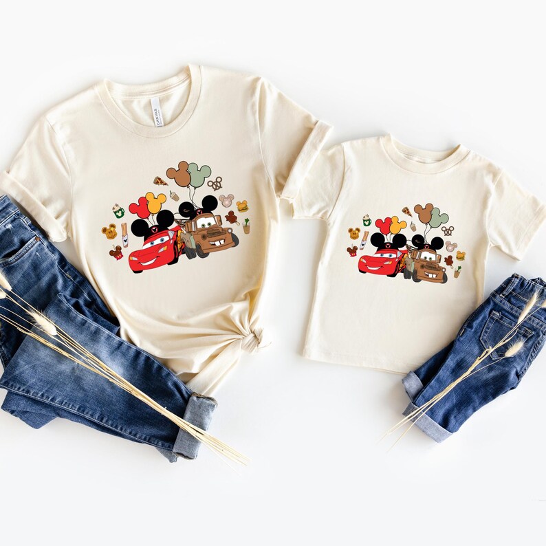 May include: Two cream-coloured t-shirts, one adult and one child size, featuring a colourful graphic of Lightning McQueen and Mater from the film Cars, with Mickey Mouse ears and balloons. The shirts are paired with blue denim jeans.