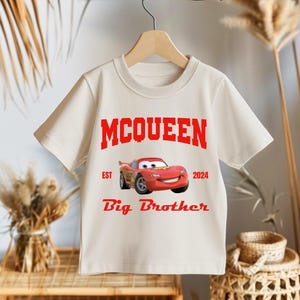 May include: Cream-colored short-sleeve t-shirt featuring the word "MCQUEEN" in red block letters above a cartoon image of a red race car. The text "EST 2024" and "Big Brother" are also in red. The shirt is hanging on a wooden hanger.