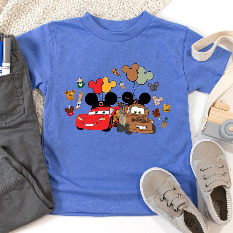 May include: A blue short-sleeved t-shirt with a graphic of Lightning McQueen and Mater from the Cars film, alongside Mickey Mouse balloons and food icons. The shirt is shown with grey trousers and trainers.