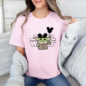 May include: Pink t-shirt featuring a green character wearing a Mickey Mouse hat and holding a Mickey Mouse balloon. The text "STAR WARS" is printed on the shirt.