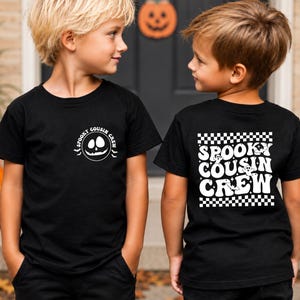 May include: A black short-sleeve t-shirt featuring a white graphic on the front left chest that says "SPOOKY COUSIN CREW" with a pumpkin face. The back displays a large white "SPOOKY COUSIN CREW" text design with ghosts and a checkerboard pattern.