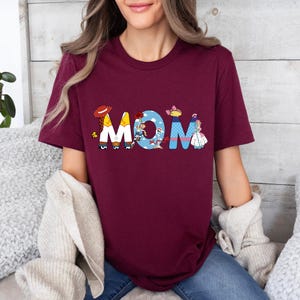 K&ouml;nnte beinhalten: Burgunderfarbenes T-Shirt mit einem Aufdruck des Wortes "MOM", das aus Figuren aus den Toy Story-Filmen besteht. Die Figuren sind Woody, Buzz Lightyear, Jessie und Bo Peep.