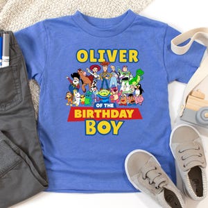 May include: A blue short-sleeved t-shirt featuring a colorful graphic of characters from a popular animated movie. The shirt has the name "Oliver" above the phrase "Birthday Boy". Gray pants and sneakers are next to the shirt.