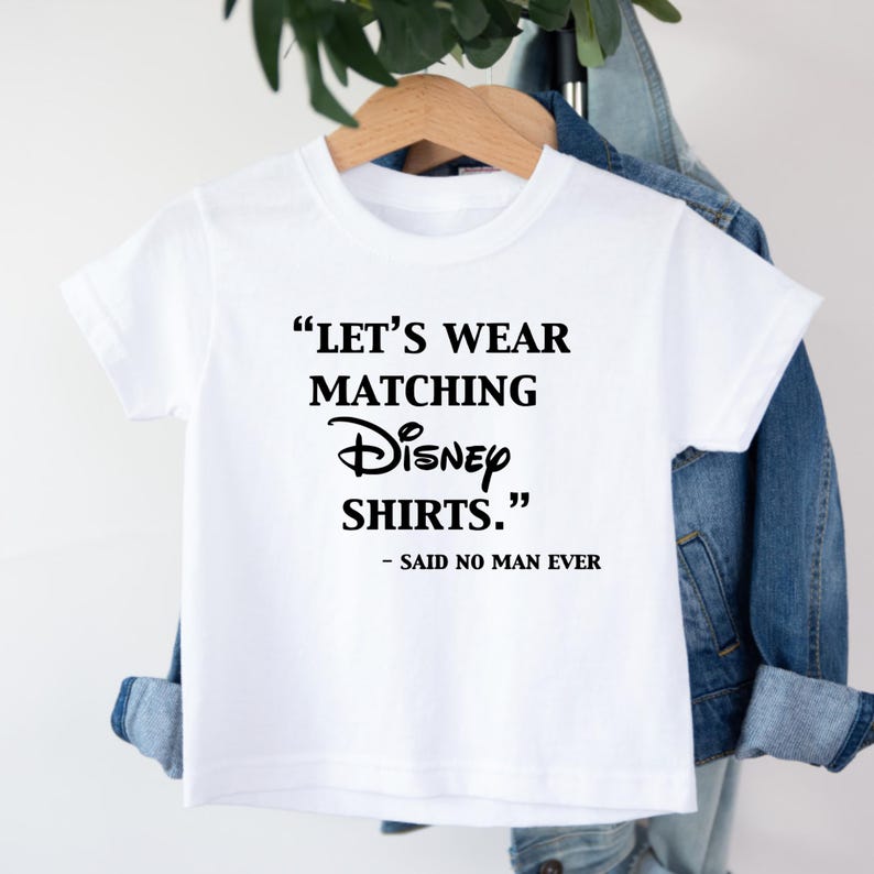 Peut inclure: Un t-shirt blanc avec le texte "LET'S WEAR MATCHING Disney SHIRTS." en noir, avec "- SAID NO MAN EVER" en dessous. Le t-shirt est suspendu &agrave; un cintre en bois.