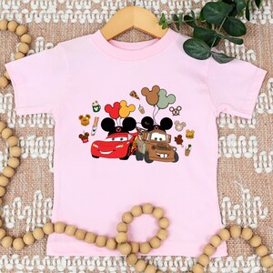 May include: A pale pink t-shirt with a cartoon graphic of Lightning McQueen and Mater from the Cars film, each with Mickey Mouse ears and balloons. Various cartoon food and character icons are also included.