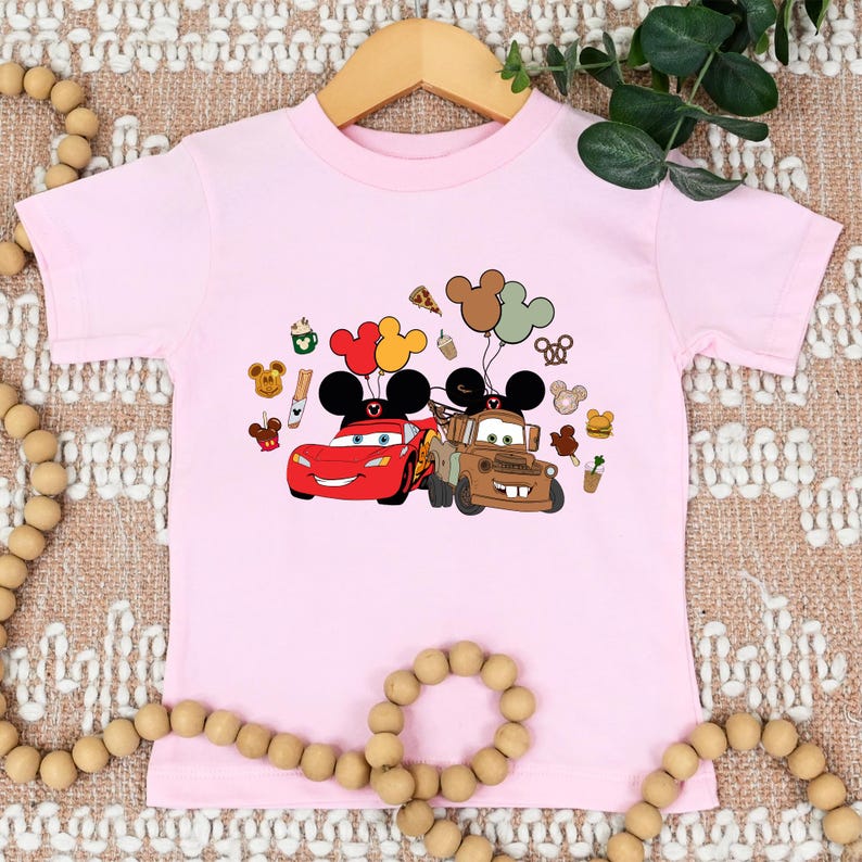 May include: A pale pink t-shirt with a cartoon graphic of Lightning McQueen and Mater from the Cars film, each with Mickey Mouse ears and balloons. Various cartoon food and character icons are also included.