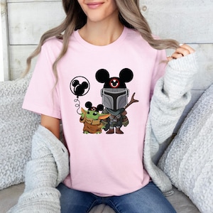 May include: Pink t-shirt with a graphic of a Mandalorian character wearing Mickey Mouse ears and holding a balloon. A small green character with large ears is standing next to him.